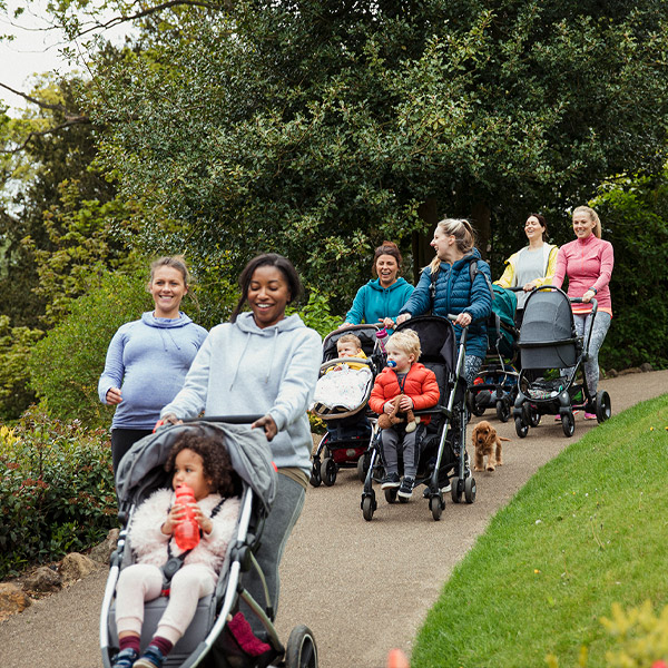 Six women walking along a park path pushing buggies and chatting