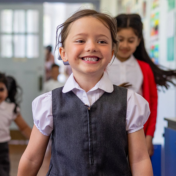 A young girl in the foreground, smiling in her school uniform in a school corridor