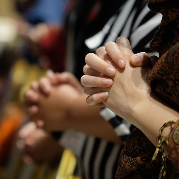 In the foreground we see an woman holding her hands together praying, in the background other people doing the same